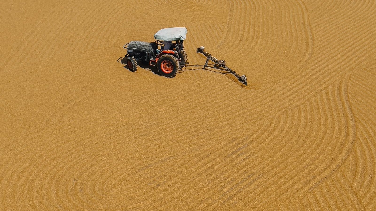 Traktor med harv drar jämna spår genom sandunderlaget på en stor ridbana, sett uppifrån